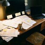 A desk covered in stacked printed documents, sticky notes, and a closed laptop under a single desk lamp, empty chair pulled back, rest of the room in shadow, no person visible.