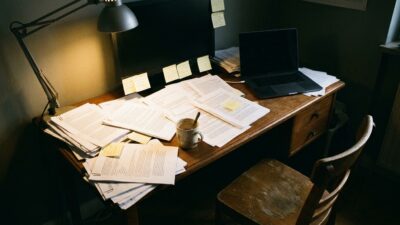 A desk covered in stacked printed documents, sticky notes, and a closed laptop under a single desk lamp, empty chair pulled back, rest of the room in shadow, no person visible.