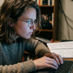 A woman with glasses sits at a desk with an open laptop, hands resting on the keyboard without typing, expression tense, a handwritten notebook open beside the computer.