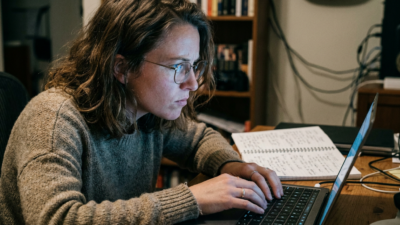 A woman with glasses sits at a desk with an open laptop, hands resting on the keyboard without typing, expression tense, a handwritten notebook open beside the computer.
