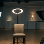 An empty host chair sits under bright ring lights on a QVC broadcast studio floor, facing a camera and teleprompter between takes.