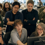 Newsroom journalists gather at desks reviewing documents and screens in a busy editorial office under natural light.